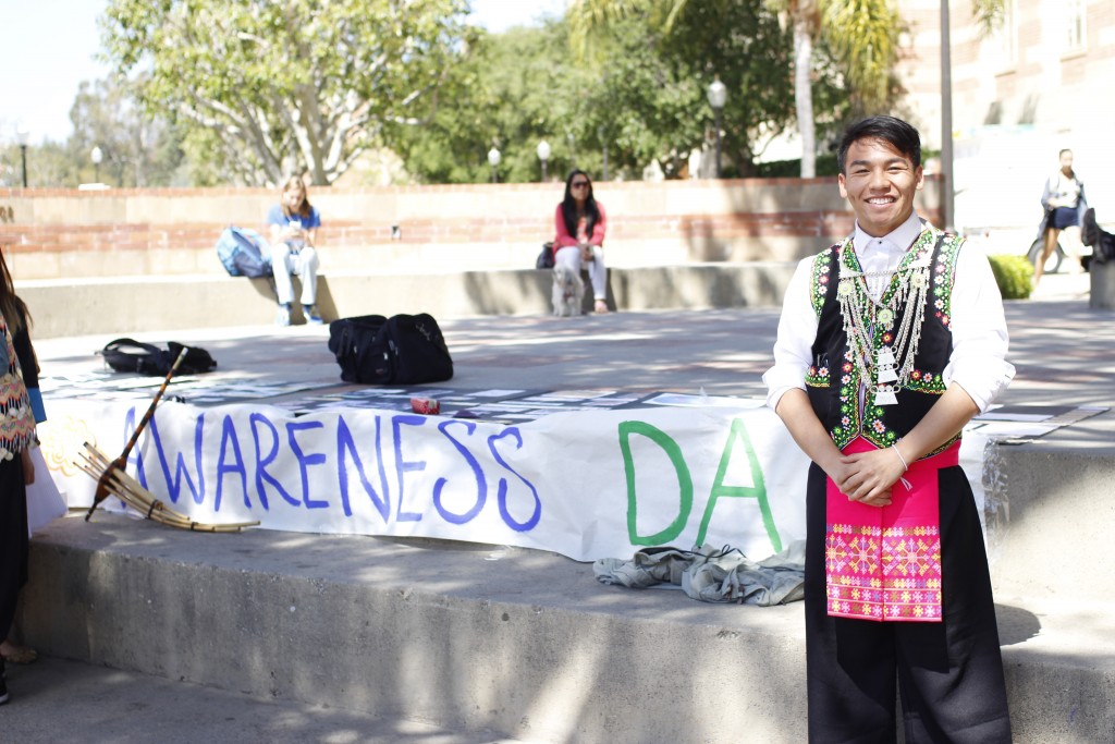 Teddy Vang poses in Hmong clothing next to a banner on McClure Stage in Bruin Plaza